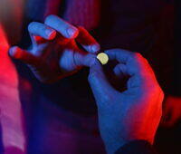 Hands holding a small yellow pill, likely GHB, in a dimly lit nightclub environment with red and blue lighting