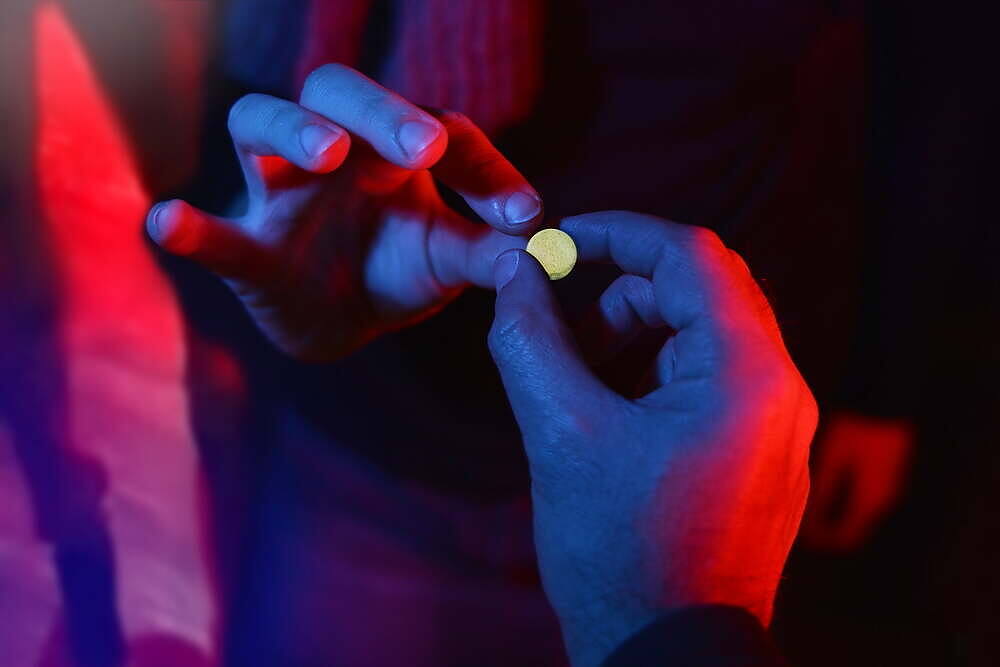 Hands holding a small yellow pill, likely GHB, in a dimly lit nightclub environment with red and blue lighting