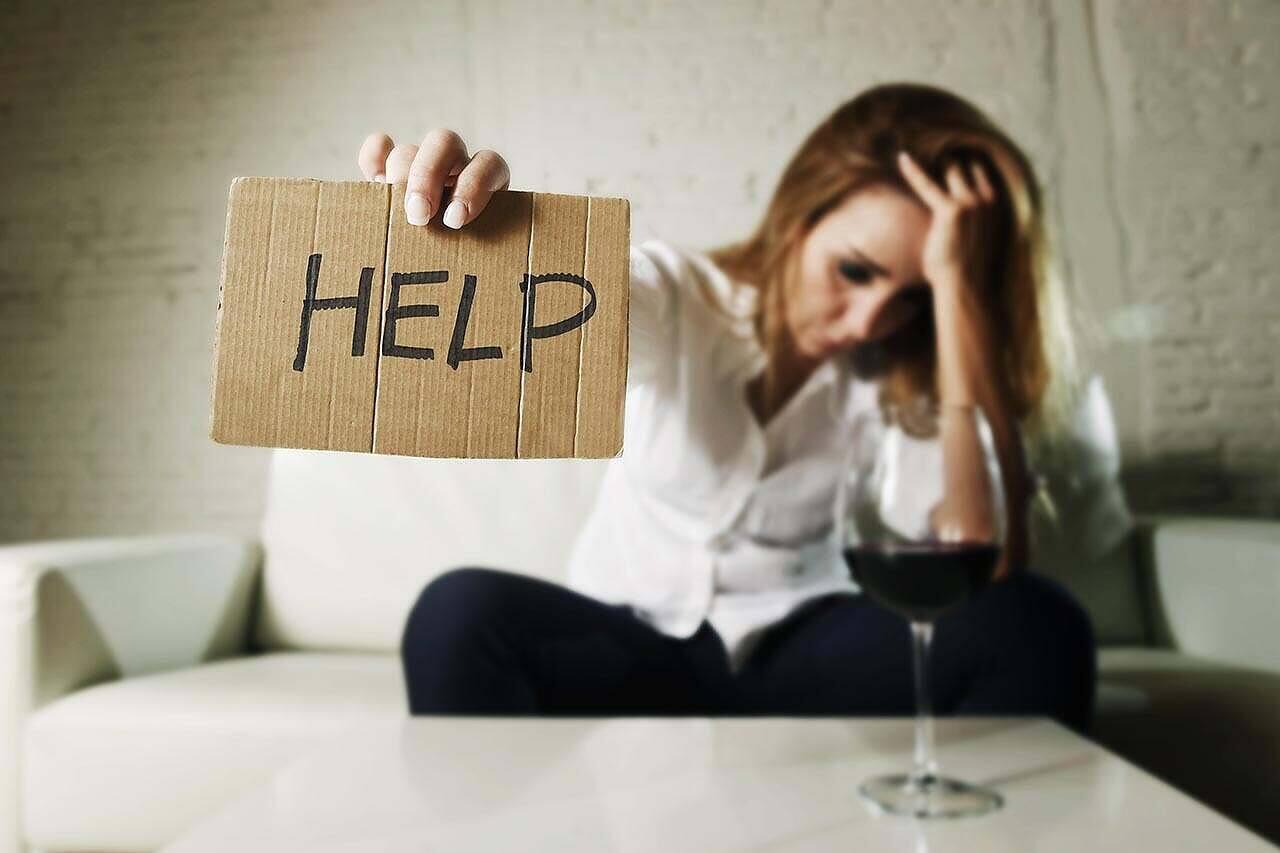 Distressed woman holding 'HELP' sign next to wine glass, illustrating struggle with alcoholism