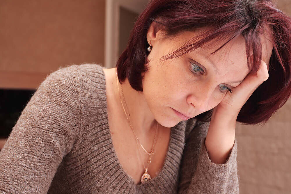 Middle-aged woman with red hair looking distressed, holding her head in her hand, depicting signs of anxiety or depression