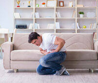 Young man kneeling by couch clutching stomach in pain, in living room with bookshelves