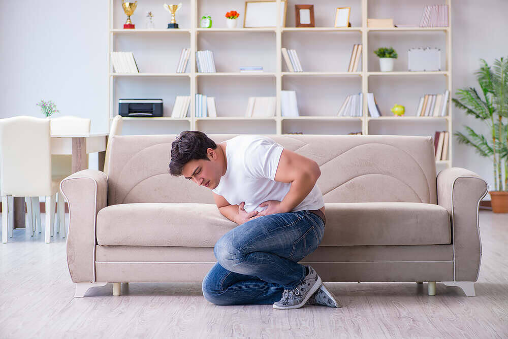 Young man kneeling by couch clutching stomach in pain, in living room with bookshelves