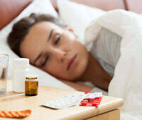 A woman asleep in bed with various prescription pill bottles and blister packs on the nightstand, illustrating potential prescription drug dependence.