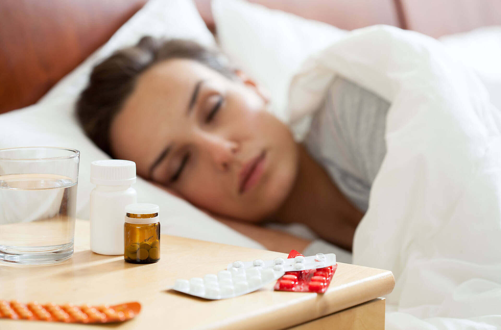 A woman asleep in bed with various prescription pill bottles and blister packs on the nightstand, illustrating potential prescription drug dependence.