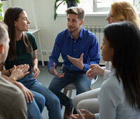 Diverse group of people engaged in a therapy session, sitting in a circle and discussing in a bright, plant-filled room