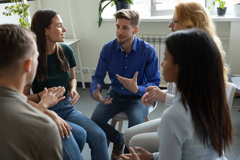 Diverse group of people engaged in a therapy session, sitting in a circle and discussing in a bright, plant-filled room