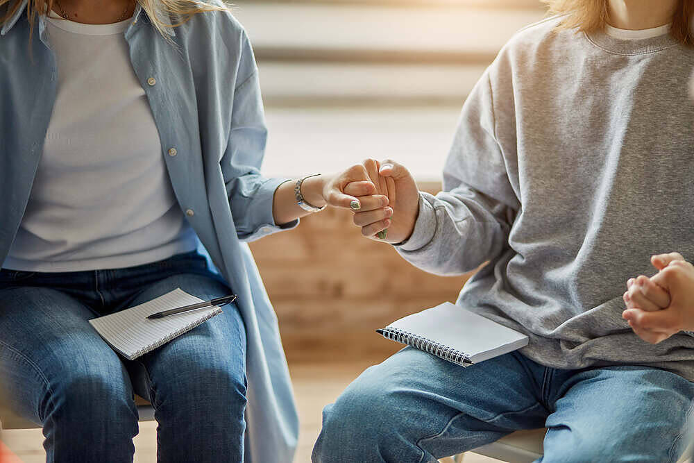 Two people holding hands during a support group meeting at a Manchester rehabilitation center, with notepads visible