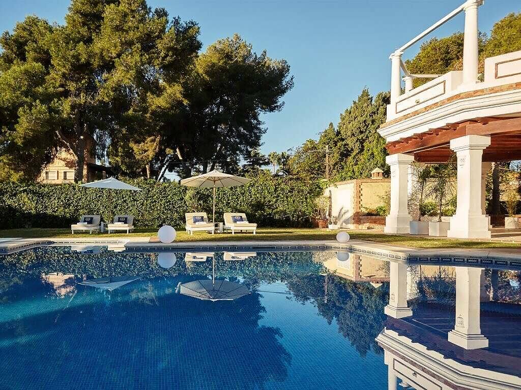 Serene swimming pool surrounded by lounge chairs and umbrellas at a luxury rehab resort, with elegant white columns and lush greenery