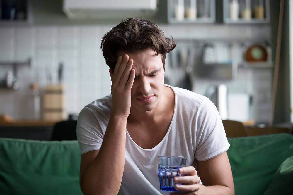 A man holding his head in pain while sitting on a couch, experiencing an alcohol-induced headache, and holding a glass of water