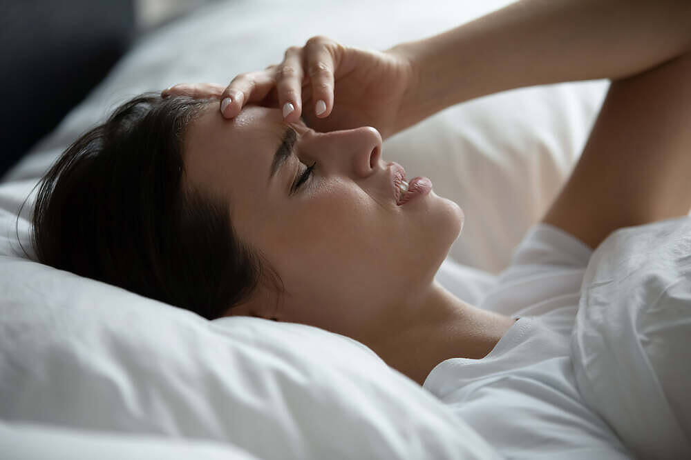 Woman lying in bed with eyes closed, touching her forehead, experiencing discomfort likely from an alcohol-related headache