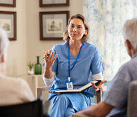 Female nurse in blue scrubs explaining to elderly couple in a care home setting, possibly discussing medication or treatment options.