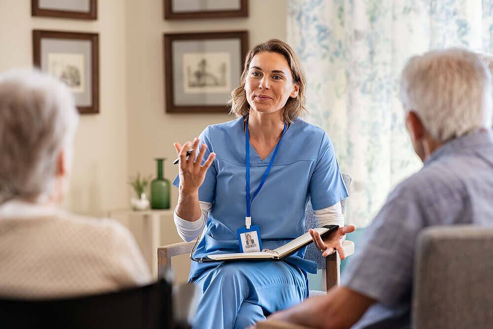 Female nurse in blue scrubs explaining to elderly couple in a care home setting, possibly discussing medication or treatment options.