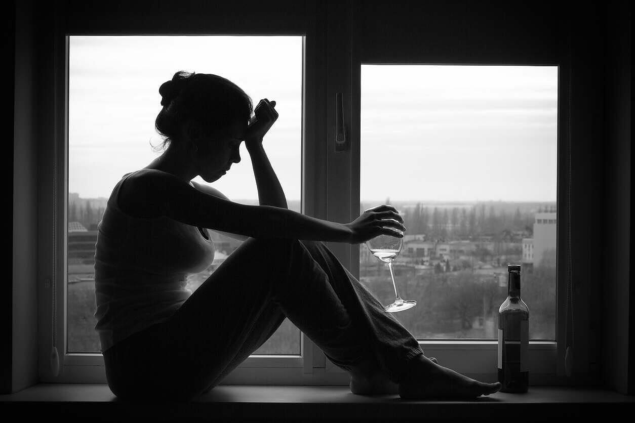 Silhouette of a woman sitting alone on a windowsill, holding a glass of wine, with a bottle nearby, illustrating the solitary nature of addiction