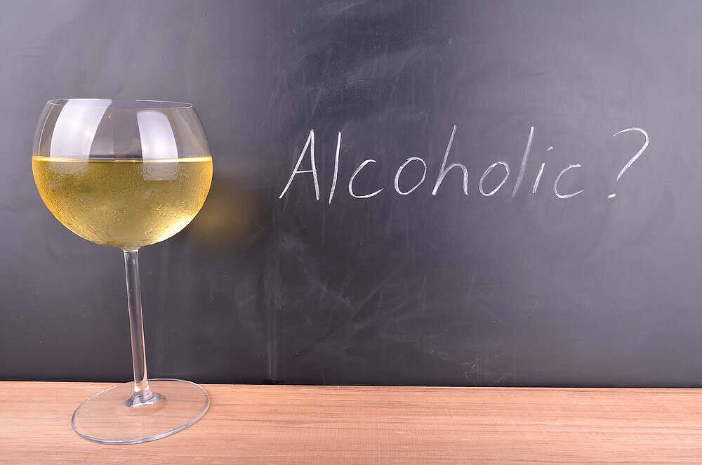 A glass of white wine next to a chalkboard with 'Alcoholic?' written on it, suggesting self-reflection on drinking habits