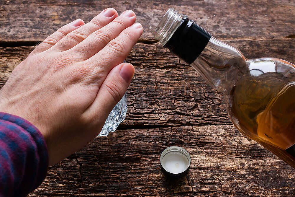 Hand gesture refusing alcohol, blocking whiskey bottle on wooden table, symbolizing quitting drinking