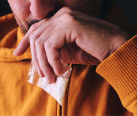 Close-up of a man in an orange sweater holding a small plastic bag containing white powder, likely cocaine, illustrating dangerous drug use