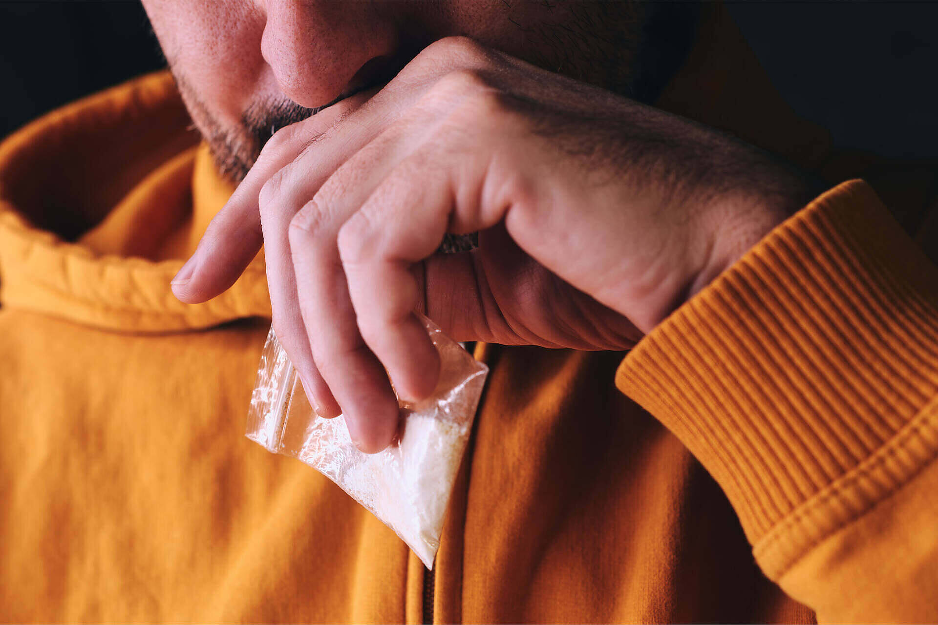 Close-up of a man in an orange sweater holding a small plastic bag containing white powder, likely cocaine, illustrating dangerous drug use