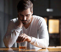 Troubled man leaning on counter, looking pensively at glass of alcohol, considering addiction treatment