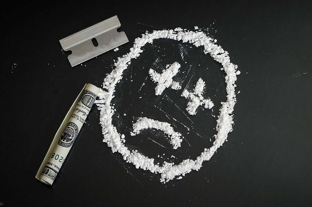 Symbolic image of cocaine addiction: sad face made from white powder, with razor blade and dollar bill