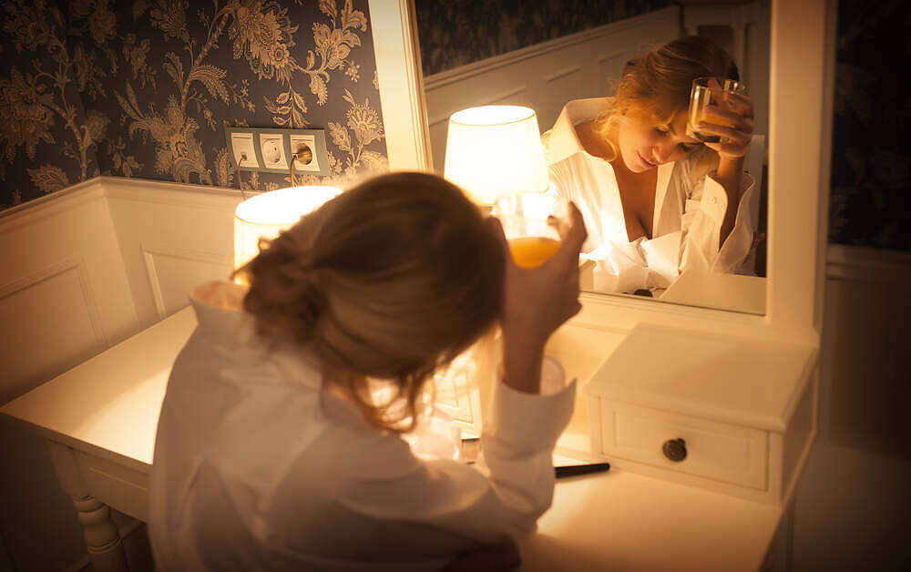 Woman in white robe drinking alcohol alone at vanity mirror, illustrating struggle with alcohol cravings