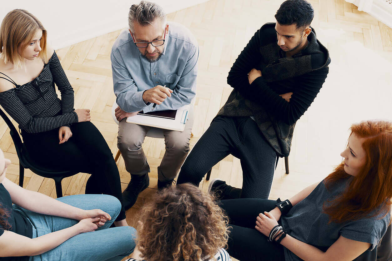 Diverse group of people sitting in a circle during a therapy or support group session, with a therapist taking notes