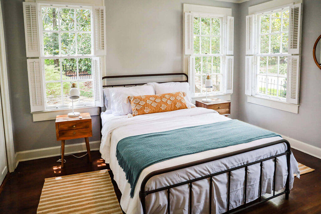 Bright, comfortable bedroom in a London alcohol rehab facility featuring a wrought iron bed, multiple windows with shutters, and a serene garden view