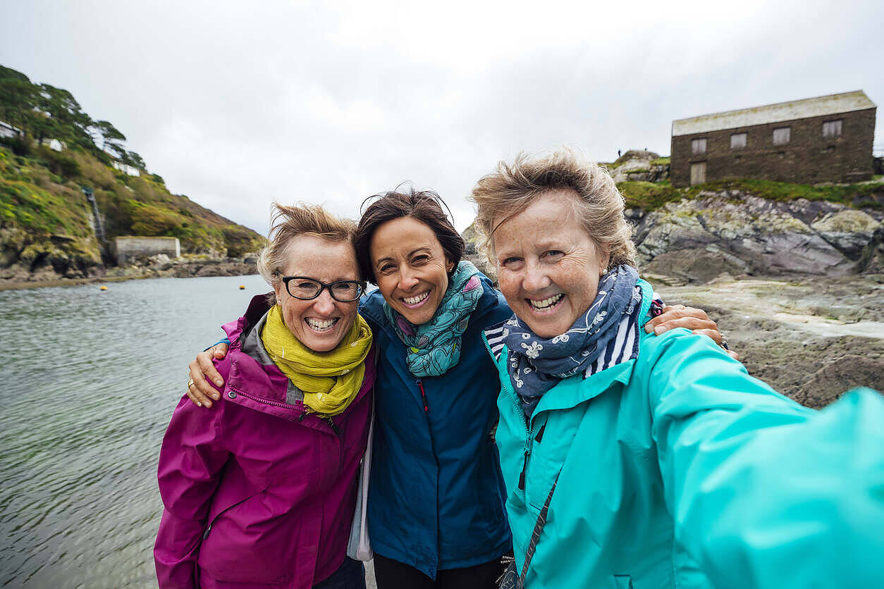 Three smiling women in colorful jackets embracing by a scenic Scottish loch, representing support and camaraderie in recovery