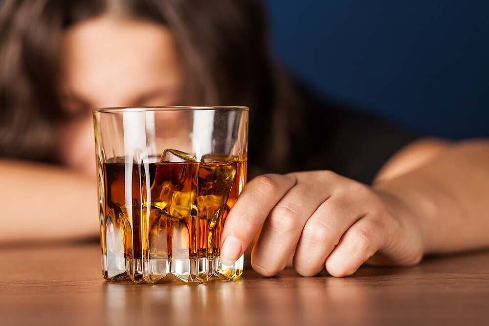 Close-up of a woman resting her head on a table next to a glass of whiskey, depicting alcohol abuse