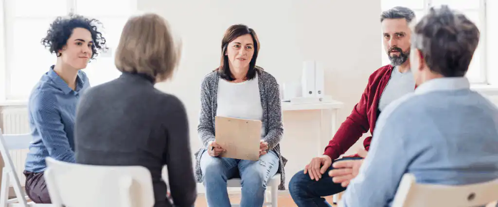 Diverse group of people sitting in a circle during a therapy session at an addiction treatment center, led by a female therapist
