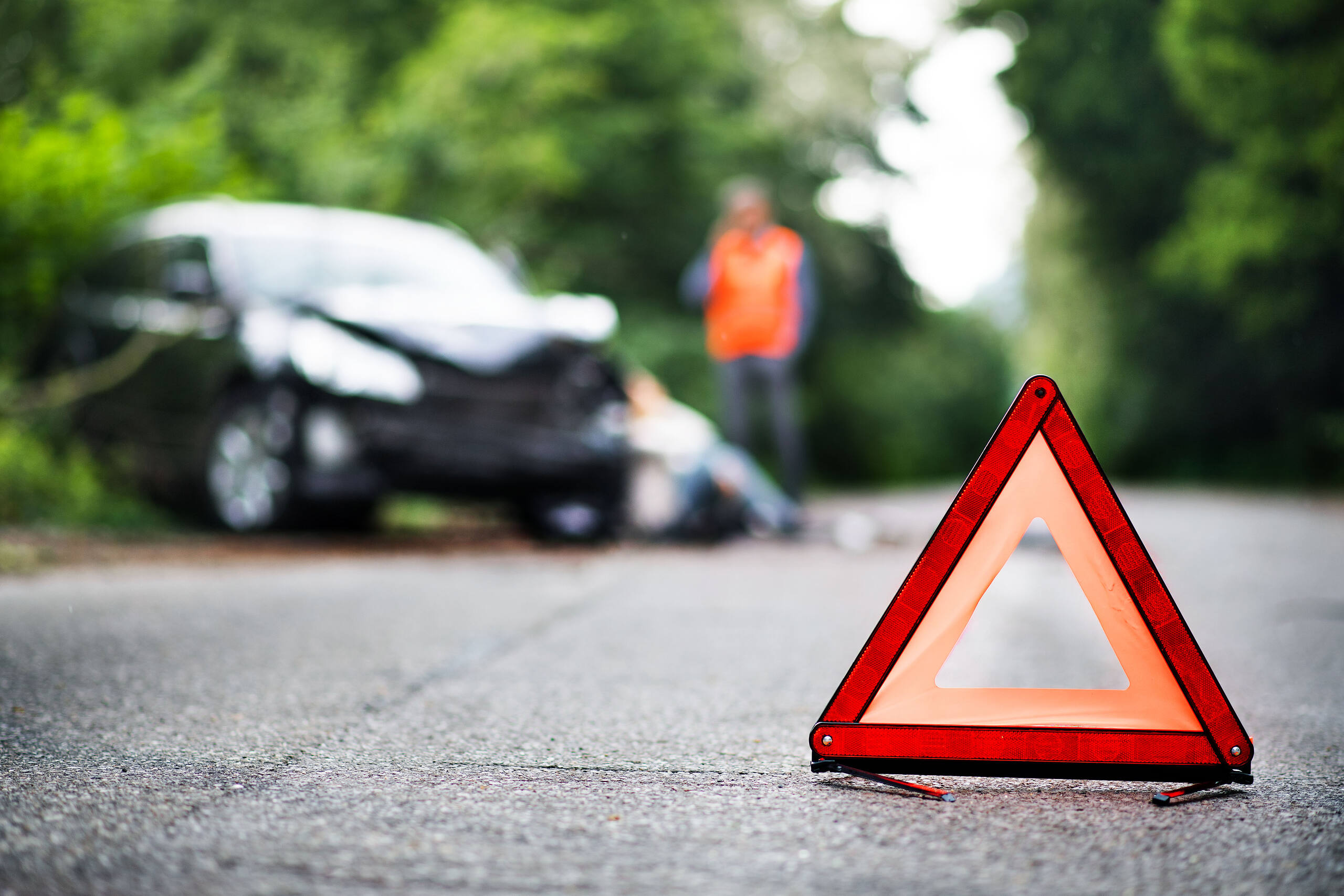 Car accident scene with warning triangle in foreground, damaged vehicle and person in high-visibility vest in background, illustrating potential consequences of driving under influence