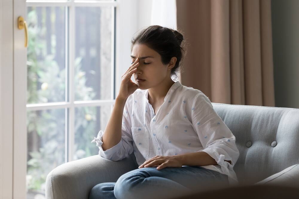 Woman experiencing discomfort and fatigue, possibly due to opiate addiction withdrawal symptoms, sitting on a couch near a window