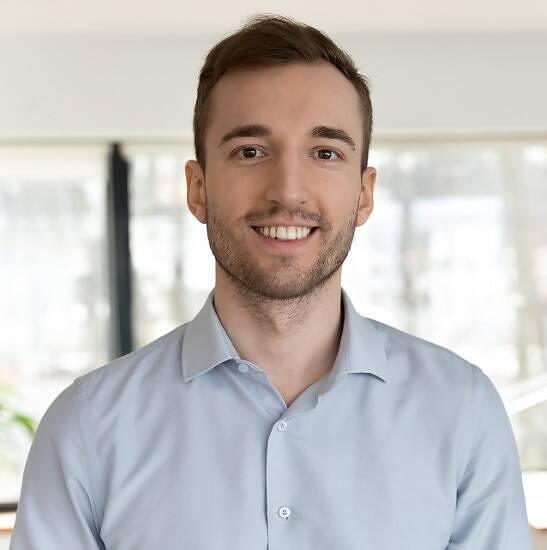 Smiling young man with short brown hair and beard in a light blue shirt, representing a recovered problem drinker in a bright office setting