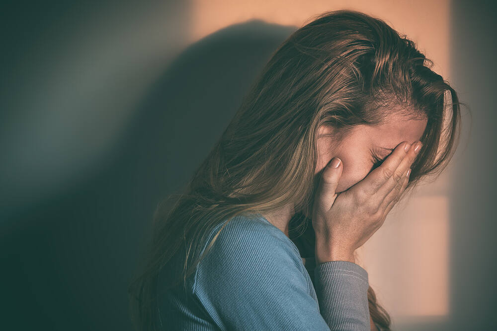 Depressed woman with long hair covering her face with hands, sitting alone in dim lighting, showing signs of emotional distress