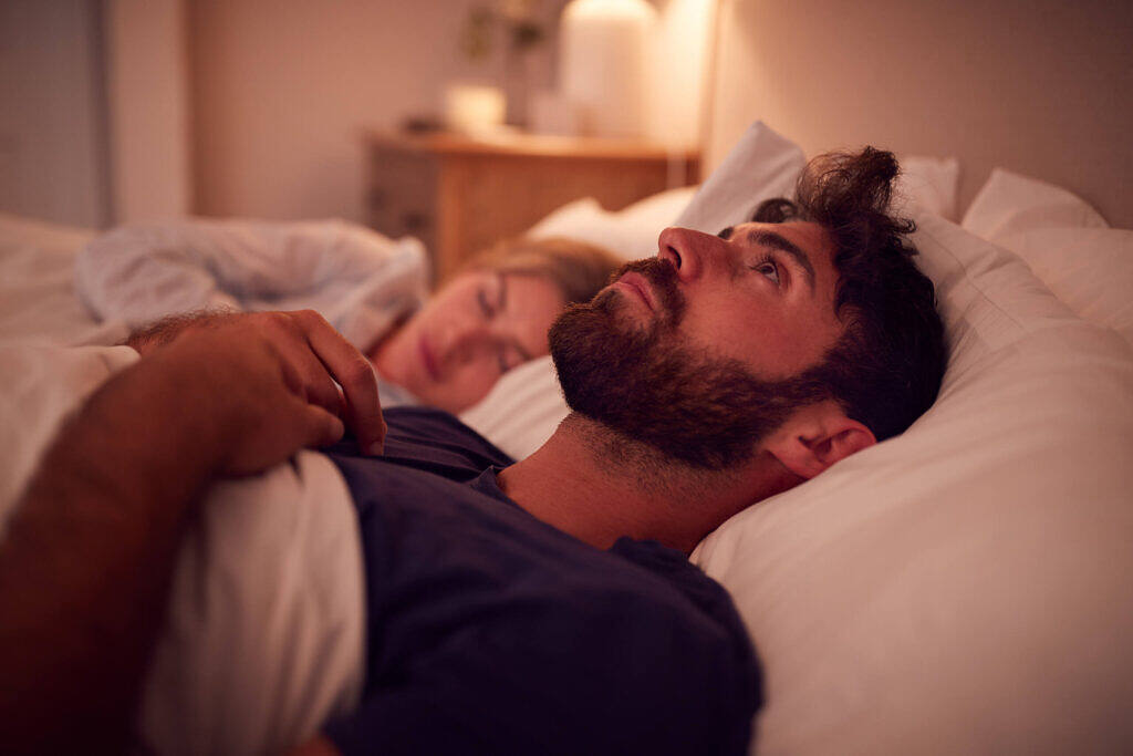 Bearded man lying awake in bed at night, partner sleeping beside him, illustrating insomnia during addiction recovery