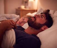 Bearded man lying awake in bed at night, partner sleeping beside him, illustrating insomnia during addiction recovery