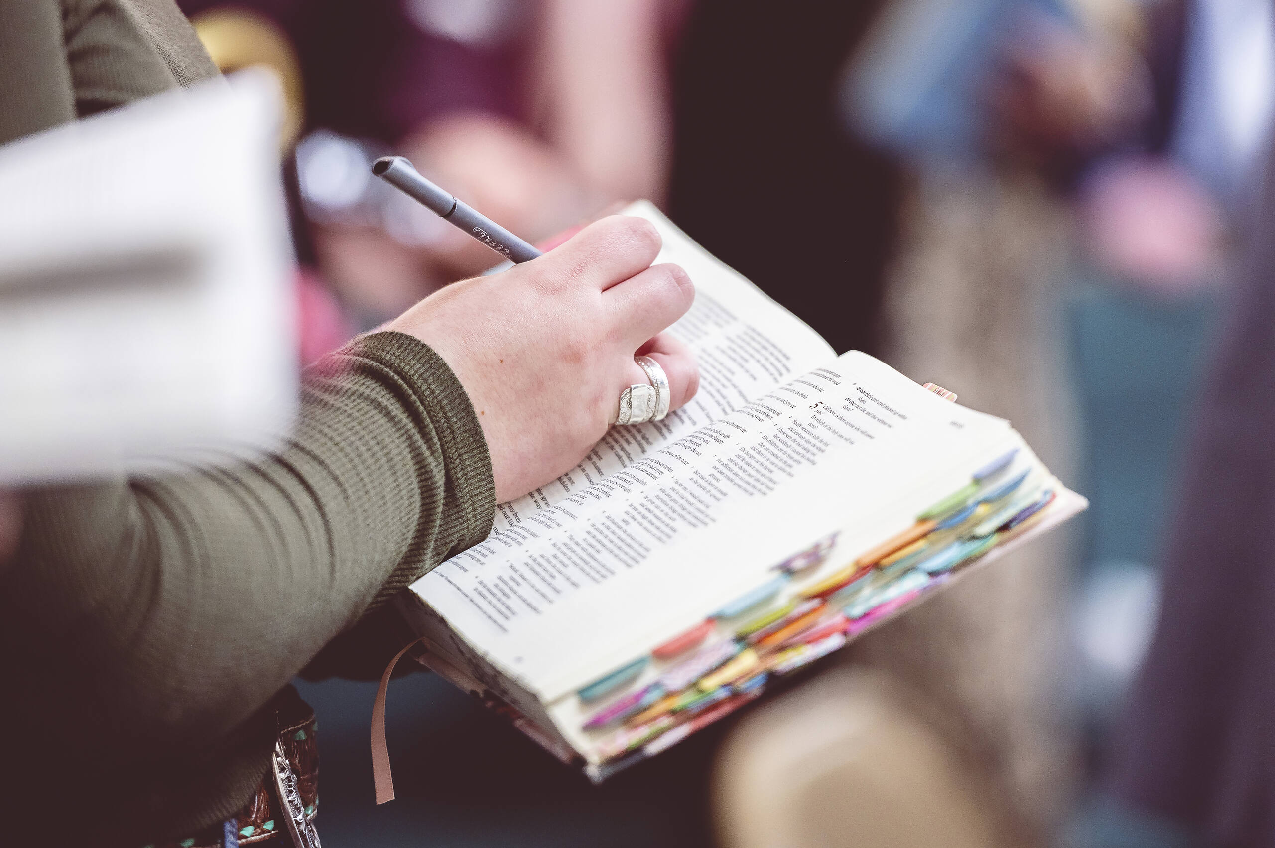 Close-up of a person's hand holding a pen over an open Bible with colorful bookmarks, illustrating Bible study in Christian rehab programs