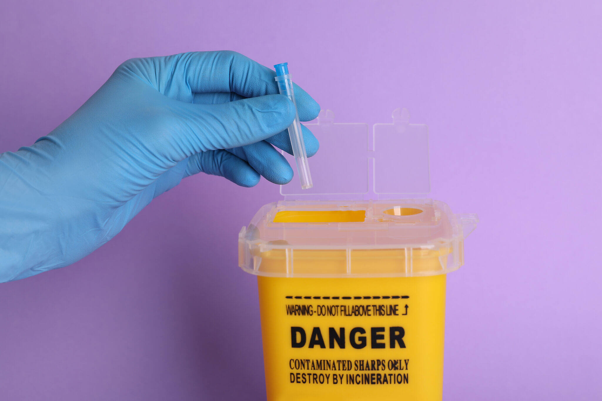 Gloved hand placing a syringe into a yellow sharps container labeled 'DANGER' against a purple background, highlighting safe disposal practices in addiction treatment