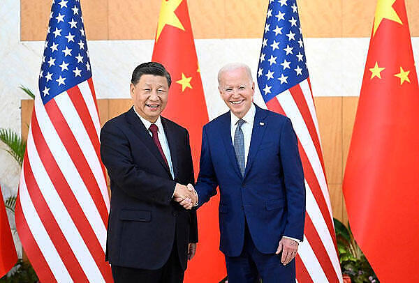 Two men in suits shaking hands: US President Joe Biden and Chinese President Xi Jinping standing in front of American and Chinese flags during a diplomatic meeting