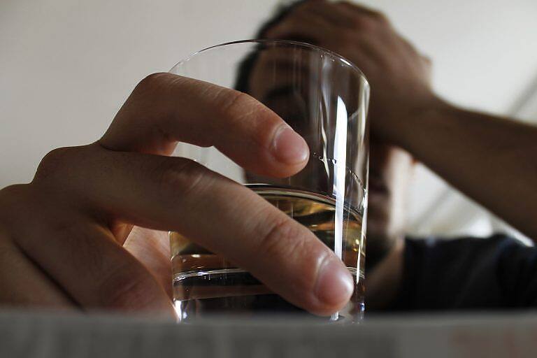 Close-up of a hand holding a glass of alcohol, with a blurred figure in the background, illustrating alcohol addiction