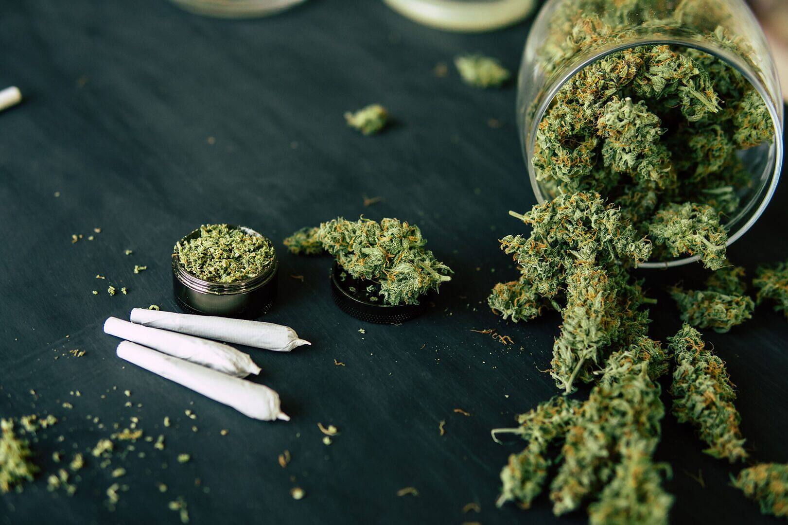 Marijuana buds spilling from a glass jar, with rolled joints and a grinder, illustrating the substances involved in cannabis addiction