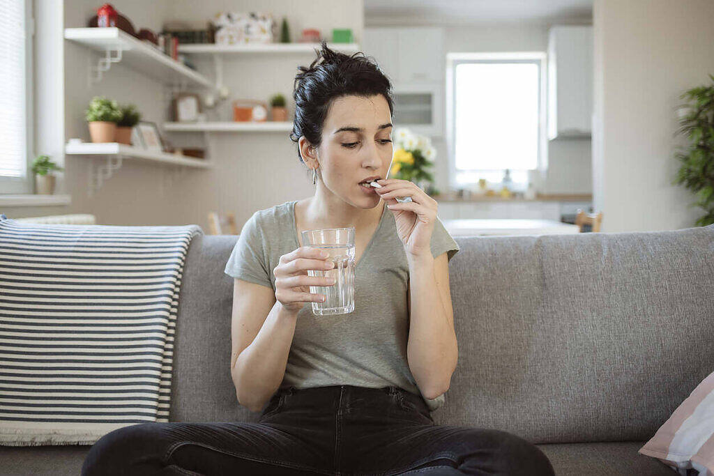 Young woman sitting on couch taking medication with glass of water in living room setting