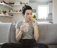 Young woman sitting on couch taking medication with glass of water in living room setting