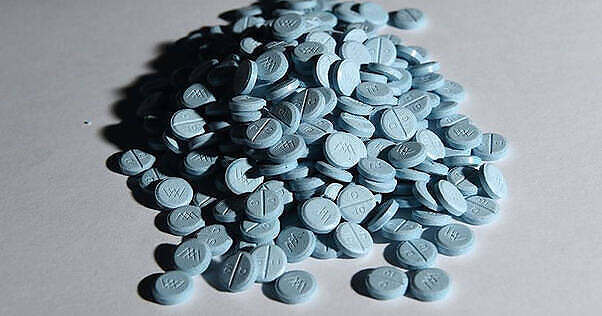 Close-up image of a large pile of light blue prescription pills, likely opioids, scattered on a white surface, illustrating the scale of prescription drug abuse