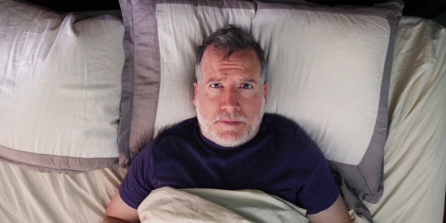 Middle-aged man with gray hair lying awake in bed, looking stressed and unable to sleep