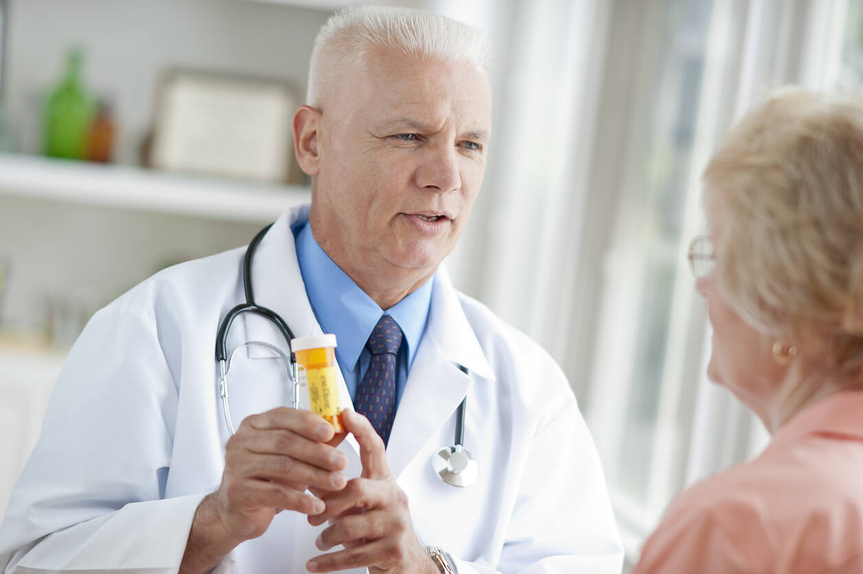 Senior male doctor in white coat holding prescription bottle, discussing medication risks with elderly female patient, emphasizing potential dangers of mixing alcohol and amoxicillin