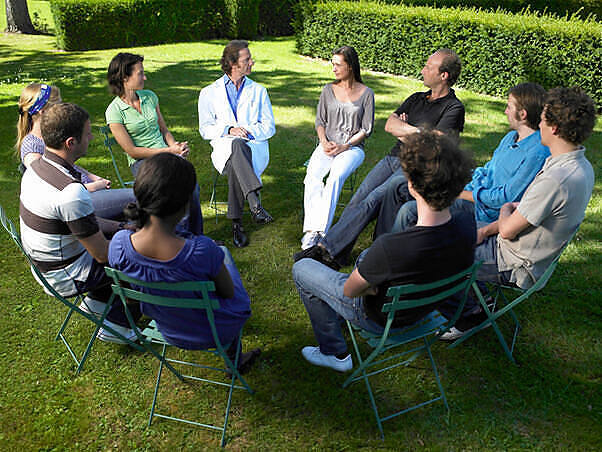 Outdoor group therapy session with diverse participants sitting in a circle, led by a therapist in a white coat, illustrating a supportive environment for addiction treatment