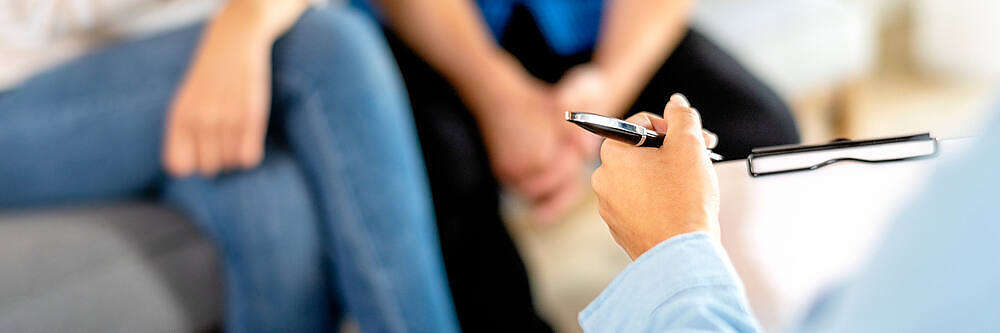 Close-up of a healthcare professional taking notes during a consultation, with a patient sitting in the background