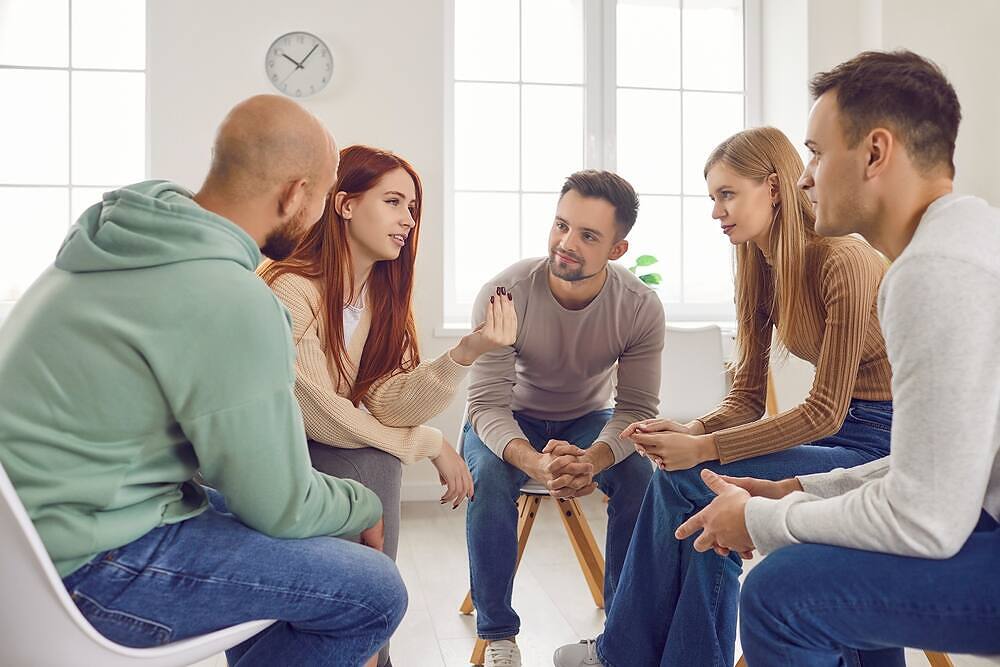 Diverse group of young adults engaged in an intimate addiction therapy session, sharing experiences and offering support in a bright, welcoming environment