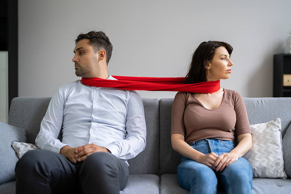 Couple sitting apart on couch, connected by red scarf, symbolizing relationship tension due to addiction