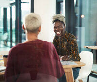 Two women, one Black wearing a headwrap and patterned top, one with short white hair, laughing together at a table in a bright office setting, depicting positive recovery support